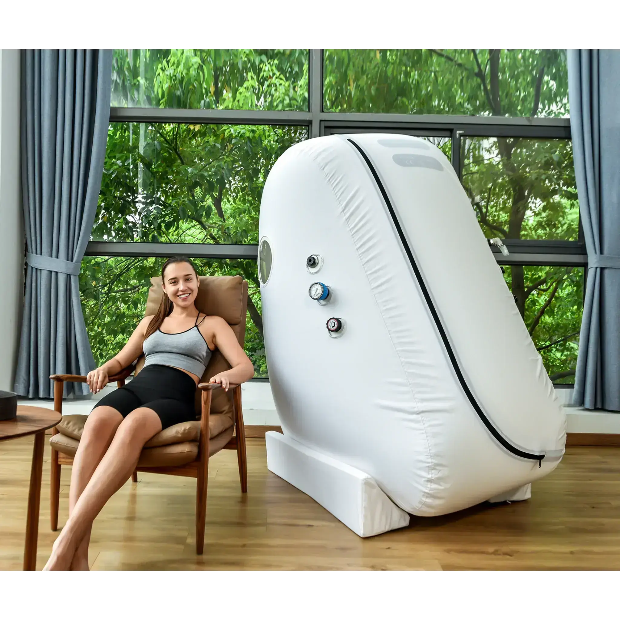 Woman comfortably seated next to Macy-Pan L1 Hyperbaric Oxygen Therapy Chamber in a bright, airy room.