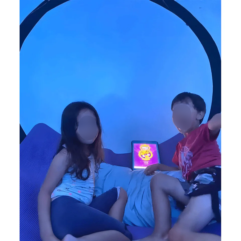 Two children relaxing inside a Newtowne 54" tent hyperbaric chamber, with a tablet displaying colorful content.